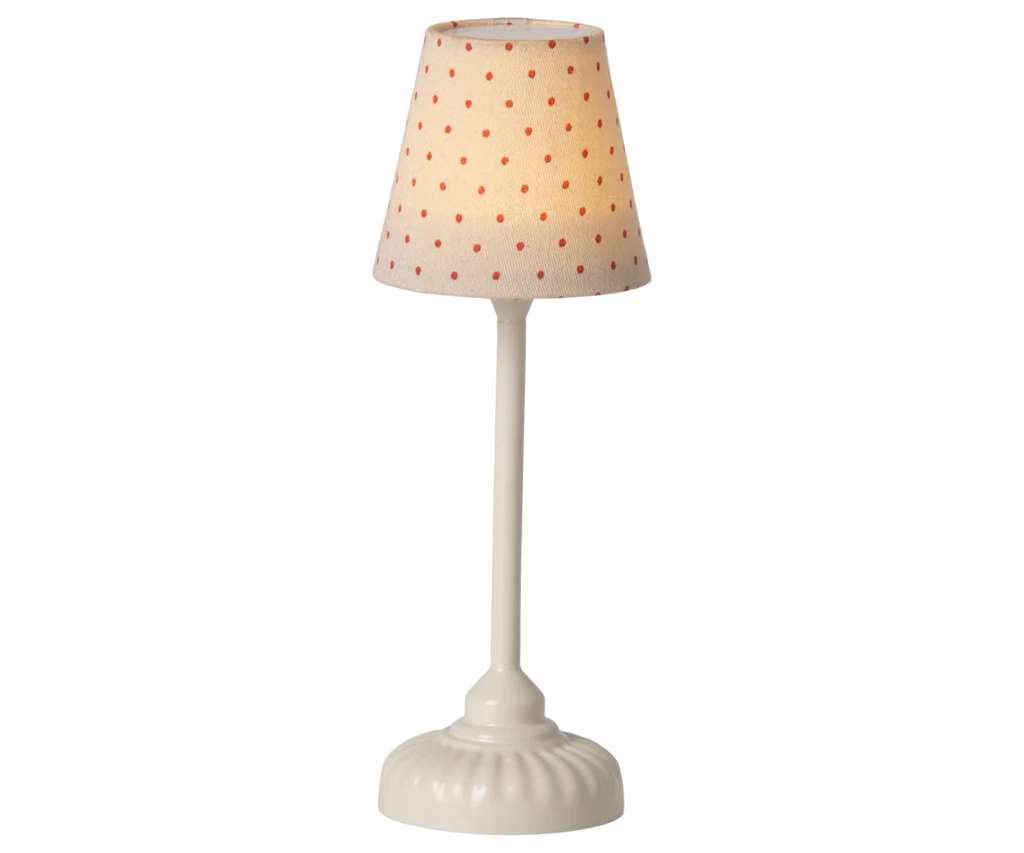 A tall white Odin Parker lamp, featuring a round, sturdy base and a beige lampshade decorated with small red polka dots, softly illuminated in the style of the Maileg Mouse Size Vintage Floor Lamp in Sand (With Battery).