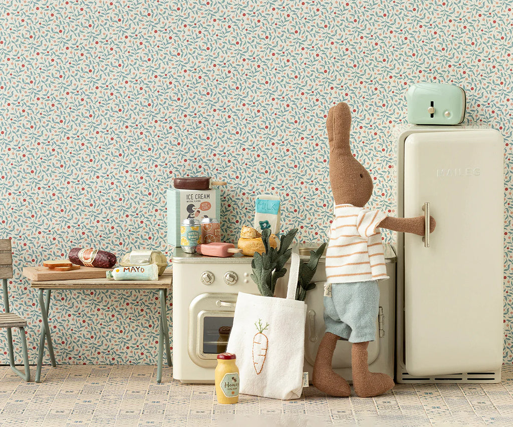 An adorable plush bunny stands next to a retro-style toy fridge, donning a striped shirt and blue shorts. The scene features a toy kitchen setup complete with the Maileg Miniature Grocery Box filled with various miniature foods, perfect for pretend play food fun, against a charming patterned wallpaper backdrop. Meet your new Maileg friend in their cozy kitchen!