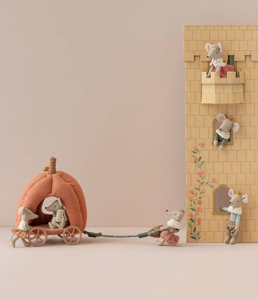 A whimsical fairytale scene with mice figures. One mouse pulls a Maileg Pumpkin Carriage carrying the royal mice family across a grey background. Nearby, a tall castle facade has other mice figures in windows and on balconies, one mouse leaning out with flowers.