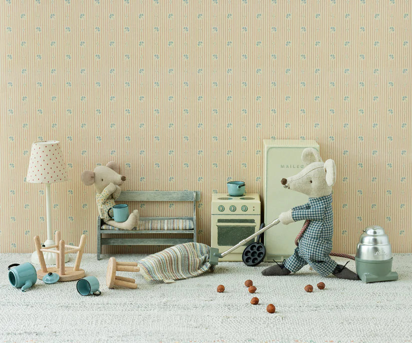 In a cozy kitchen scene adorned with vintage toys, two plush mice create a charming setting. One mouse busily vacuums beside a miniature stove on a Maileg Striped Rug, Medium, while the other relaxes on a bench holding a cup. Scattered small kitchen objects and nuts enhance the inviting ambiance against the backdrop of patterned wallpaper.