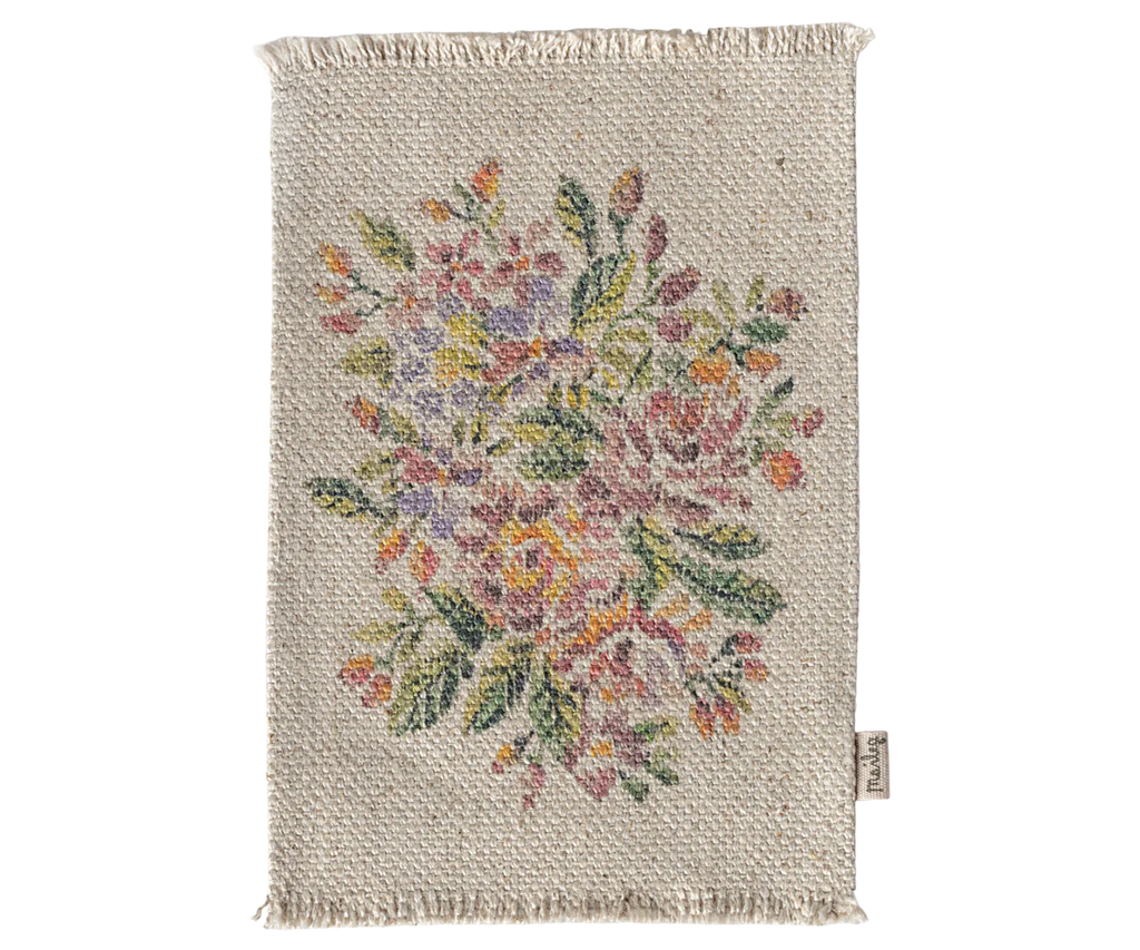 The Maileg Nursery Set includes a small rectangular woven rug with a central floral pattern, multicolored flowers, green leaves on beige, fringed edges, and a fabric tag—ideal for Maileg Miniatures or under the Maileg Mouse Closet.