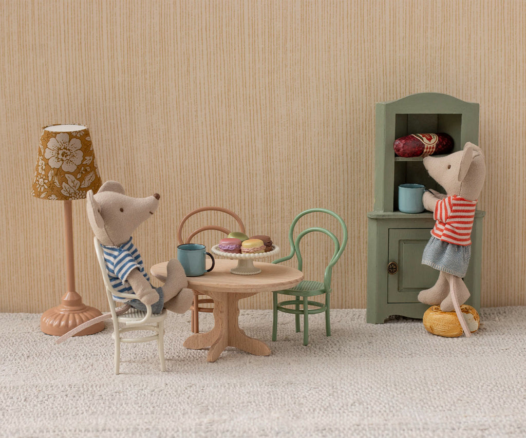Two toy mice sit at a tiny table with cups and pastries on charming dollhouse furniture. They use the Maileg Vintage Chair, Mouse Size - Dark Powder, creating a cozy scene with dishes and a floral lamp in the background.