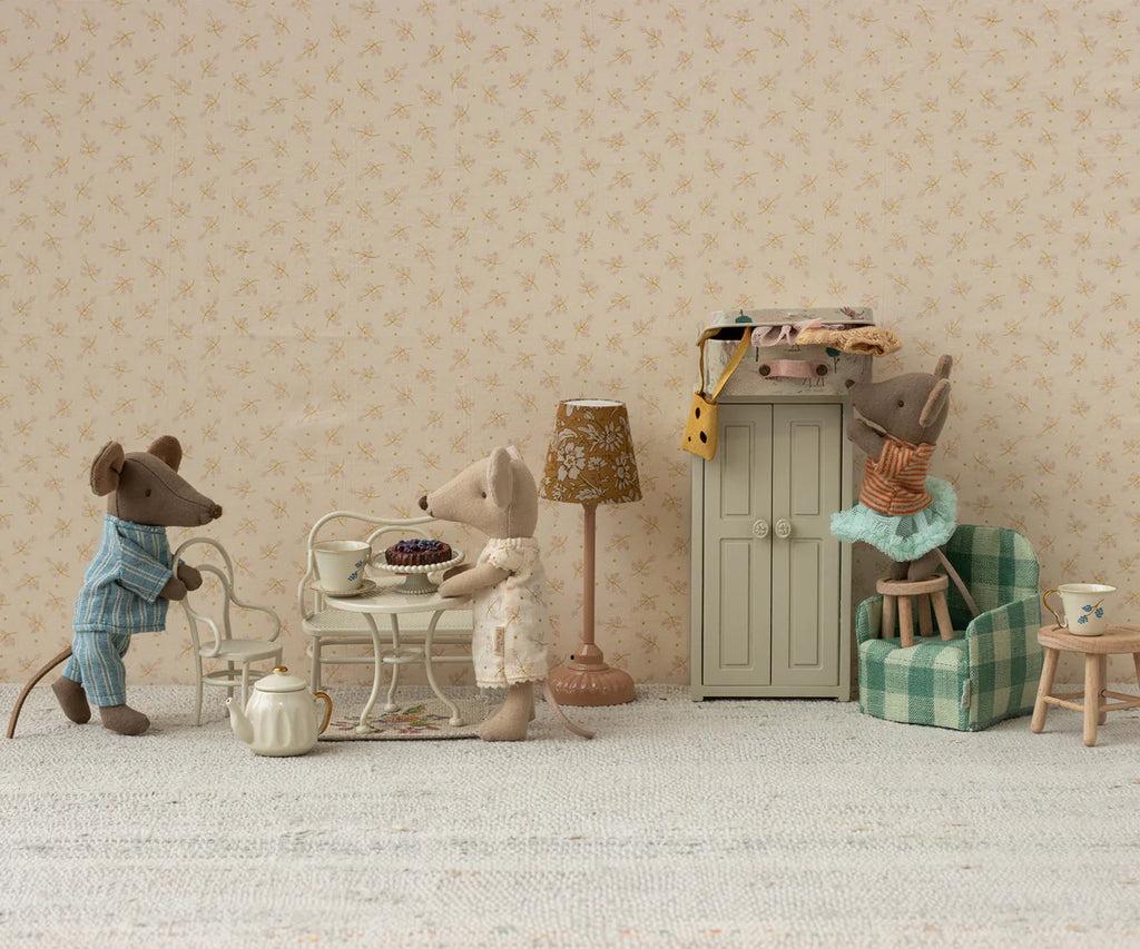 Three toy mice in various outfits have a tea party in the Maileg Mouse Café Set Table & Chair - Off-White. One serves tea while another sits with pastries, and a third reaches for a high shelf, surrounded by miniature furniture against a floral-patterned wall.