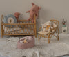 A cozy nursery scene features the Maileg Carry Cot, Large - Rose as a portable bed for plush friends, a matching rattan chair with a stuffed bunny, a basket with a soft toy, and a toy washing machine on a textured cream rug.