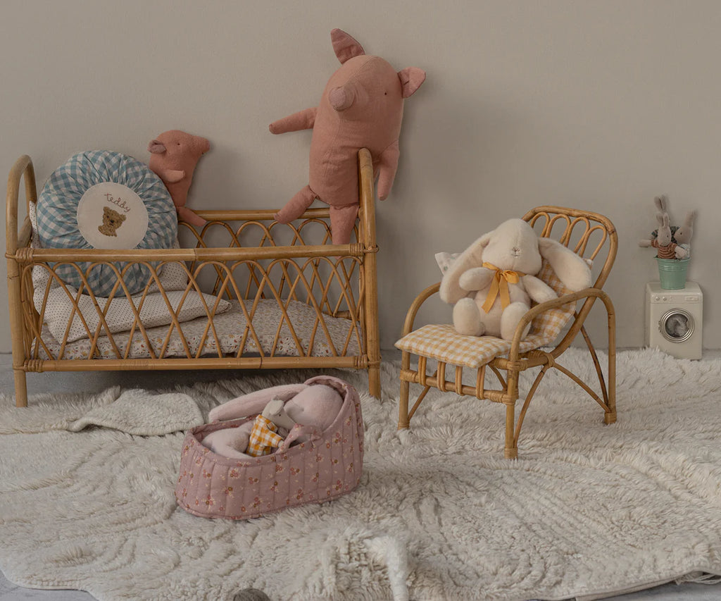 A cozy nursery scene features the Maileg Carry Cot, Large - Rose as a portable bed for plush friends, a matching rattan chair with a stuffed bunny, a basket with a soft toy, and a toy washing machine on a textured cream rug.