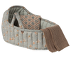 The Maileg Carry Cot, Large - Blue, is a soft quilted baby bassinet with a blue floral pattern. It serves as a cozy portable bed and includes a folded patterned blanket and a knitted brown blanket draped over the side.
