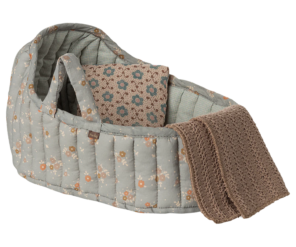 The Maileg Carry Cot, Large - Blue, is a soft quilted baby bassinet with a blue floral pattern. It serves as a cozy portable bed and includes a folded patterned blanket and a knitted brown blanket draped over the side.