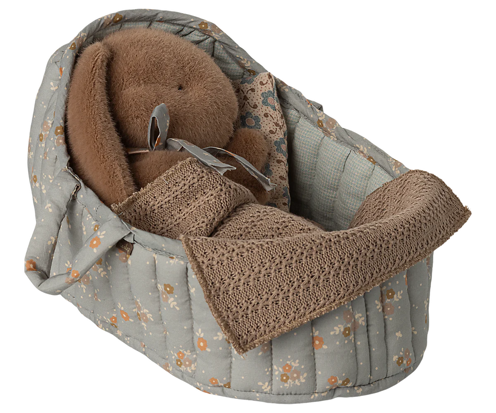 A plush brown bunny with a pacifier rests in the Maileg Carry Cot, Large - Blue, featuring a gray floral pattern and a cozy knitted blanket.