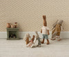 Five pastel-dressed stuffed rabbits are in a cozy room with floral wallpaper. One sits in the Maileg My Babysitter - Multi Dots chair, three smaller ones play together, and one stands nearby. A green shelf with pitchers and logs is behind them.