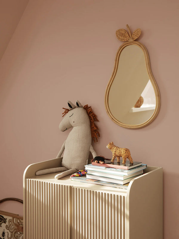 A cozy children's room corner with a cream radiator cover topped with a stuffed unicorn, a few books, and a small wooden animal toy. A whimsical Ferm Living Pear Mirror with a bamboo rattan frame.