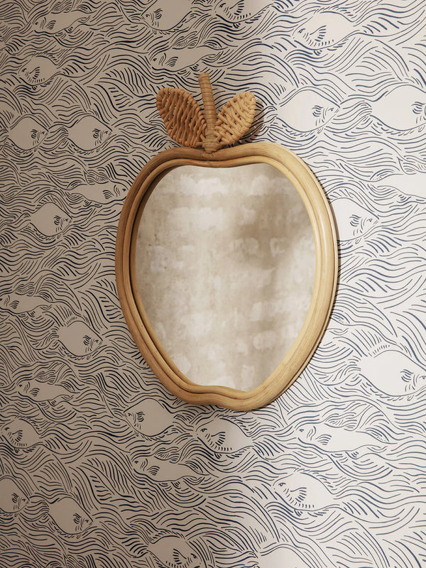 A unique Ferm Living Apple Mirror with a rattan frame mounted on a wall with whimsical fish-patterned wallpaper. The top of the mirror features a textured, decorative stalk and leaf.