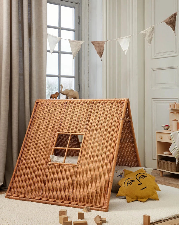 A cozy kids' room with a Ferm Living Braided Tent, inside which a cat peeks out. Decor includes a playful cushion, toy blocks, and a string of pennants. Light streams