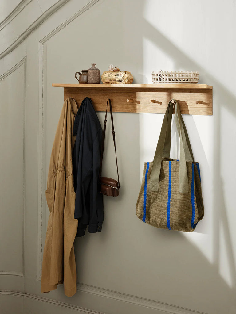 A neatly organized Ferm Living Large Place Rack - Oak with two drawers, displaying a brown jug, a small basket, and a test tube rack. Below, coats hang on hooks beside a tote bag and a shoulder bag.