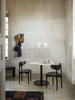 A minimal dining area features a round white table with black base, two black chairs, a modern white pendant lamp, and a Ferm Living Place Rack - Small displaying kitchenware on a light-painted wall. Natural light filters in from a nearby window.