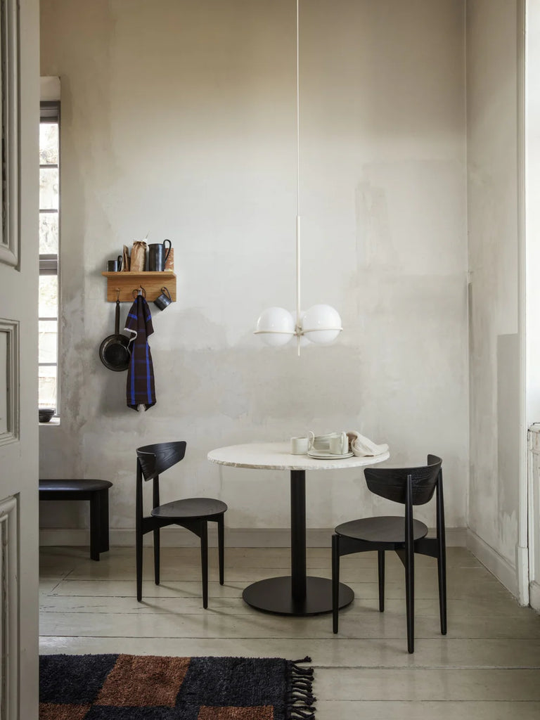 A minimal dining area features a round white table with black base, two black chairs, a modern white pendant lamp, and a Ferm Living Place Rack - Small displaying kitchenware on a light-painted wall. Natural light filters in from a nearby window.