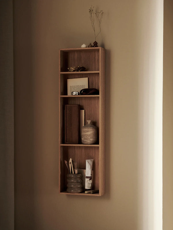 A vertical, wall-mounted Ferm Living Bon Shelf - Oiled Oak with various aesthetic storage solutions including books, a vase with dried branches, and small storage containers, illuminated by soft light from the right.