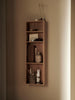A vertical, wall-mounted Ferm Living Bon Shelf - Oiled Oak with various aesthetic storage solutions including books, a vase with dried branches, and small storage containers, illuminated by soft light from the right.