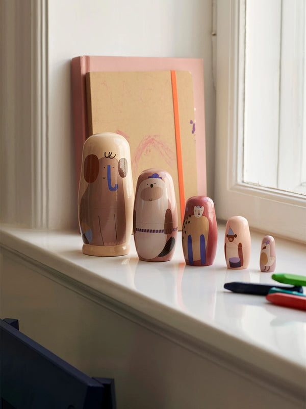 A set of nested Ferm Living Critter Nesting Dolls featuring cute animal designs sits on a sunlit windowsill, accompanied by a large sketchbook and colored pencils.