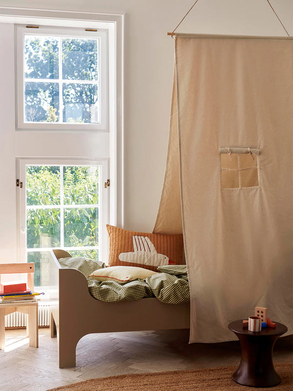 A cozy children's bedroom with a Ferm Living Settle Bed Canopy - Off-White covered in green checkered GOTS certified organic cotton bedding, a large curtain partition, a round table with building blocks, and large windows showing greenery