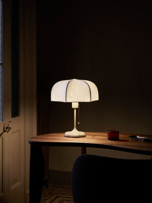 The Poem Table Lamp - White/Cashmere features a translucent shade and pull chain switch, elegantly resting on a wooden table in a dimly lit room. A small red container and closed notebook enhance the Baroque ambiance, while part of a chair is visible in the foreground.