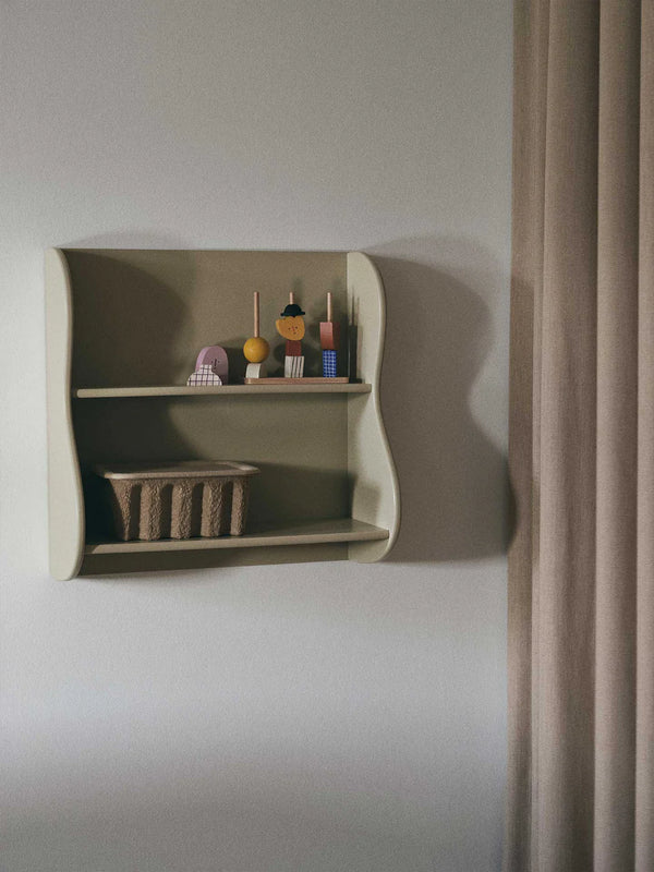 A minimalist beige Ferm Living Slope Shelf - Cashmere displaying a collection of decorative items next to a draped curtain in a softly lit room.