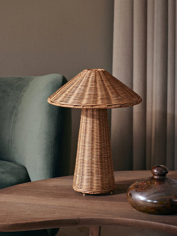 A Ferm Living Dou Table Lamp - Natural, mushroom-shaped, stands on a side table next to a green velvet chair, with a textured glass jar beside it, set against a backdrop of muted beige curtains.