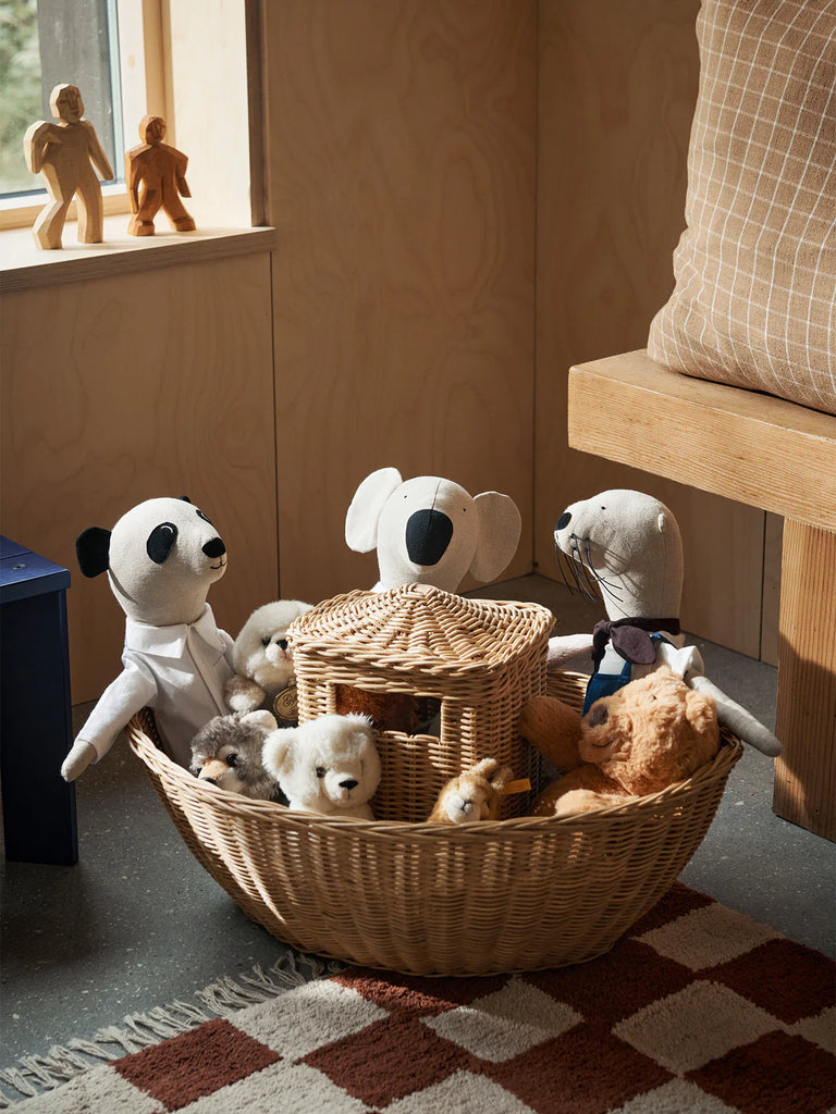 A cozy scene with a Braided Ark filled with various plush animals including a panda, dog, and teddy bear, located next to a wooden bench and under a sunlit window.