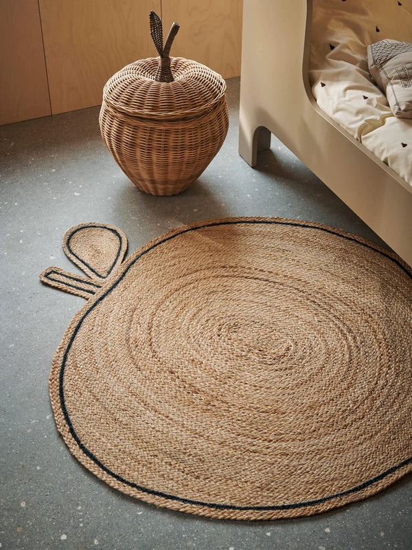 A neatly arranged interior scene featuring a large Ferm Living Apple Braided Jute Rug on the floor, a woven basket next to a low bed with beige bedding, and a playful bunny-shaped floor mat in the corner.