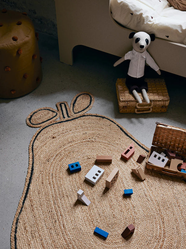 A child's bedroom floor with a Ferm Living Pear Braided Jute Rug shaped like a bee, a plush panda toy sitting on a small wicker bench, and scattered colorful wooden blocks.