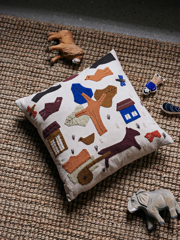 A Ferm Living Village Cushion - Off-White featuring vibrant shapes depicting animals, a house, and trees lies on a textured woven rug. This GOTS certified organic cotton cushion cover is surrounded by wooden animal figurines, including an elephant and a cow, creating a rustic and playful scene.
