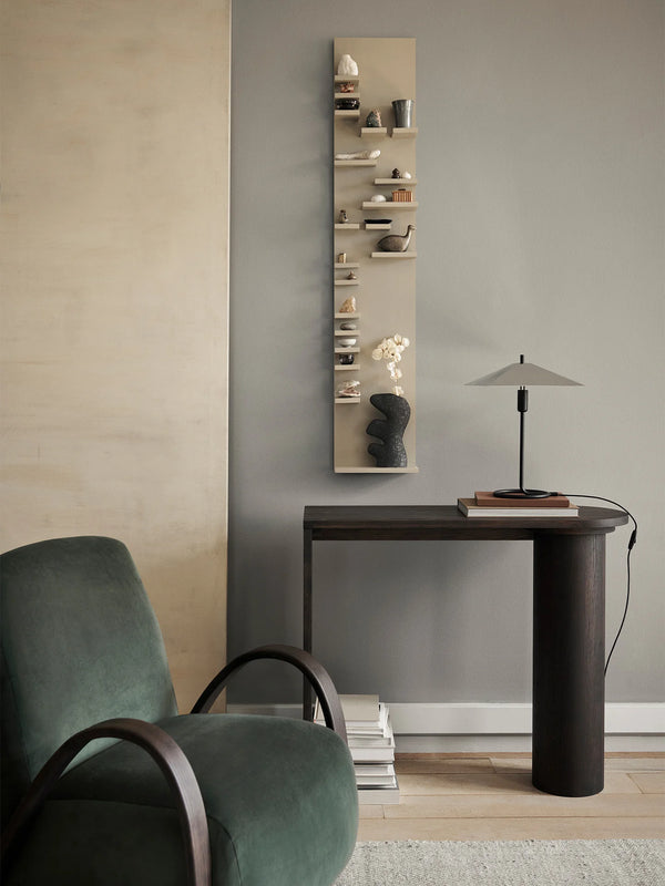 A minimalist living room featuring a green armchair, a dark wooden table with a lamp and books, and a Ferm Living Parade Shelf - Cashmere filled with small decorative items on a grey wall.