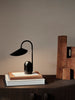 The Ferm Living Arum Portable Lamp - Black, featuring a curved neck, sits on stacked books on a dark wooden cabinet. Beside it, brown decorative wooden blocks lean against a plain beige wall.