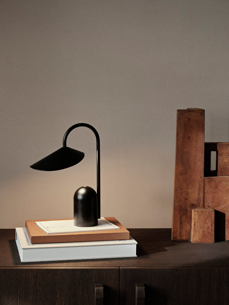 The Ferm Living Arum Portable Lamp - Black, featuring a curved neck, sits on stacked books on a dark wooden cabinet. Beside it, brown decorative wooden blocks lean against a plain beige wall.