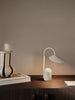 The Ferm Living Arum Portable Lamp in Cashmere, featuring a curved stem, sits on a dark wooden cabinet beside a ceramic vase and a closed book against a plain beige wall.
