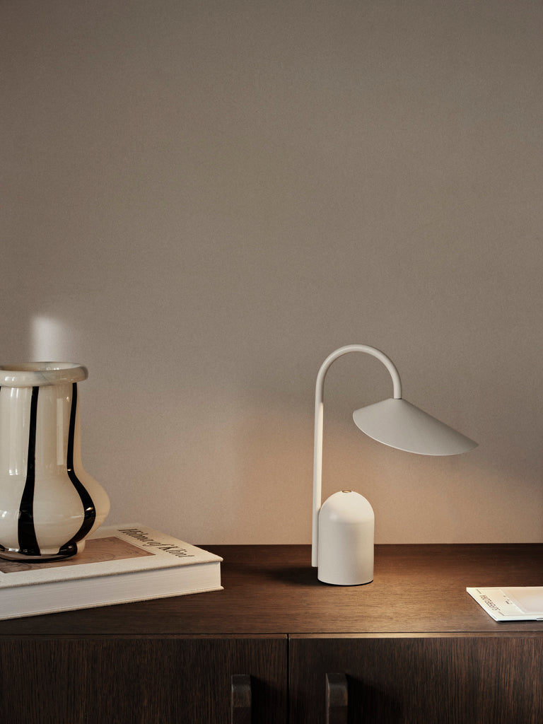 The Ferm Living Arum Portable Lamp in Cashmere, featuring a curved stem, sits on a dark wooden cabinet beside a ceramic vase and a closed book against a plain beige wall.