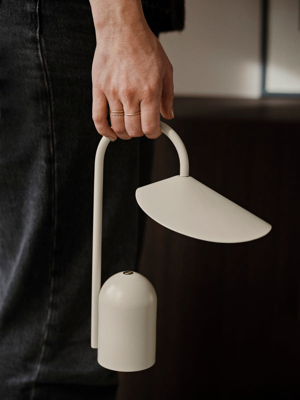A person in dark pants holds the Ferm Living Arum Portable Lamp - Cashmere, a modern LED lamp with a curved handle, touch dimmer, and angled shade, in their left hand.