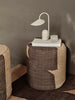 A round woven ottoman with a geometric pattern holds the Ferm Living Arum Portable Lamp in Cashmere, stacked books, and a small translucent cup against a taupe wall, creating a minimal, cozy setting.