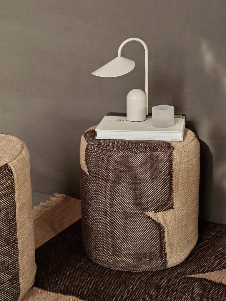A round woven ottoman with a geometric pattern holds the Ferm Living Arum Portable Lamp in Cashmere, stacked books, and a small translucent cup against a taupe wall, creating a minimal, cozy setting.