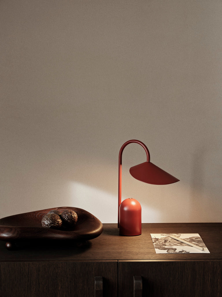 A Ferm Living Arum Portable Lamp in red sits on a dark wooden cabinet beside a sculpted wooden bowl with objects and a black-and-white print, all set against a plain beige wall.