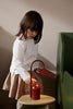 A young child with brown hair, in a white shirt and beige skirt, stands by a small table and touches the Ferm Living Arum Portable Lamp in red, illuminating the softly lit room.