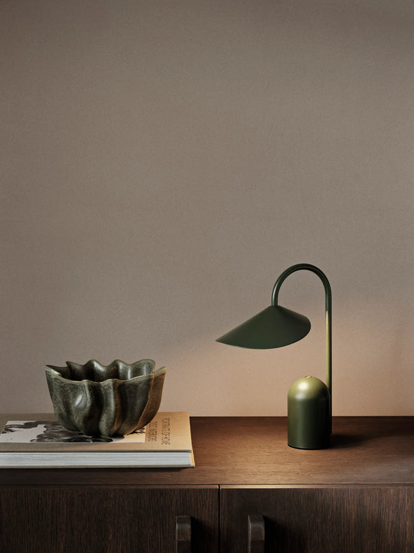 The Ferm Living Arum Portable Lamp in grass green and a textured ceramic bowl are placed on stacked books atop a dark wooden cabinet against a plain beige wall.