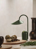 A Ferm Living Arum Portable Lamp in Grass Green sits on a countertop beside dark ceramic vases, fresh artichokes, and wooden cutting boards, creating a stylish and earthy kitchen scene.