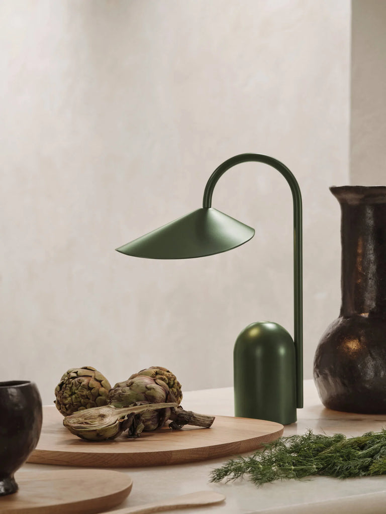 A Ferm Living Arum Portable Lamp in Grass Green sits on a countertop beside dark ceramic vases, fresh artichokes, and wooden cutting boards, creating a stylish and earthy kitchen scene.