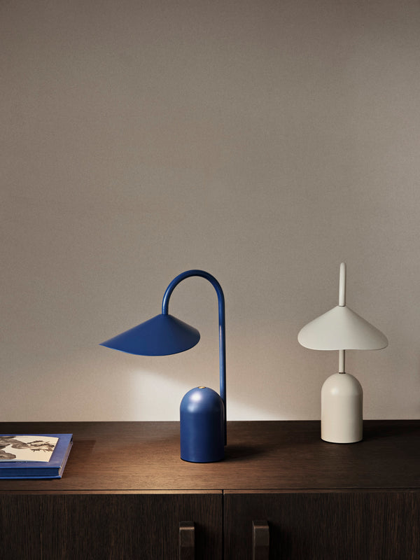 A minimalist setup featuring the Ferm Living Arum Portable Lamp in blue and a white rechargeable lamp on a dark wooden surface, beside a blue book with a black-and-white photo, set against a plain beige wall.