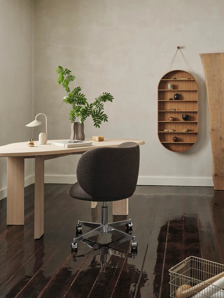 A minimalist home office features a light wood desk, black swivel chair, white lamp, and a plant in a vase. The Ferm Living Oval Oak Dorm Shelf displays small decor. Dark wood floors and a wire basket in the corner add modern storage.