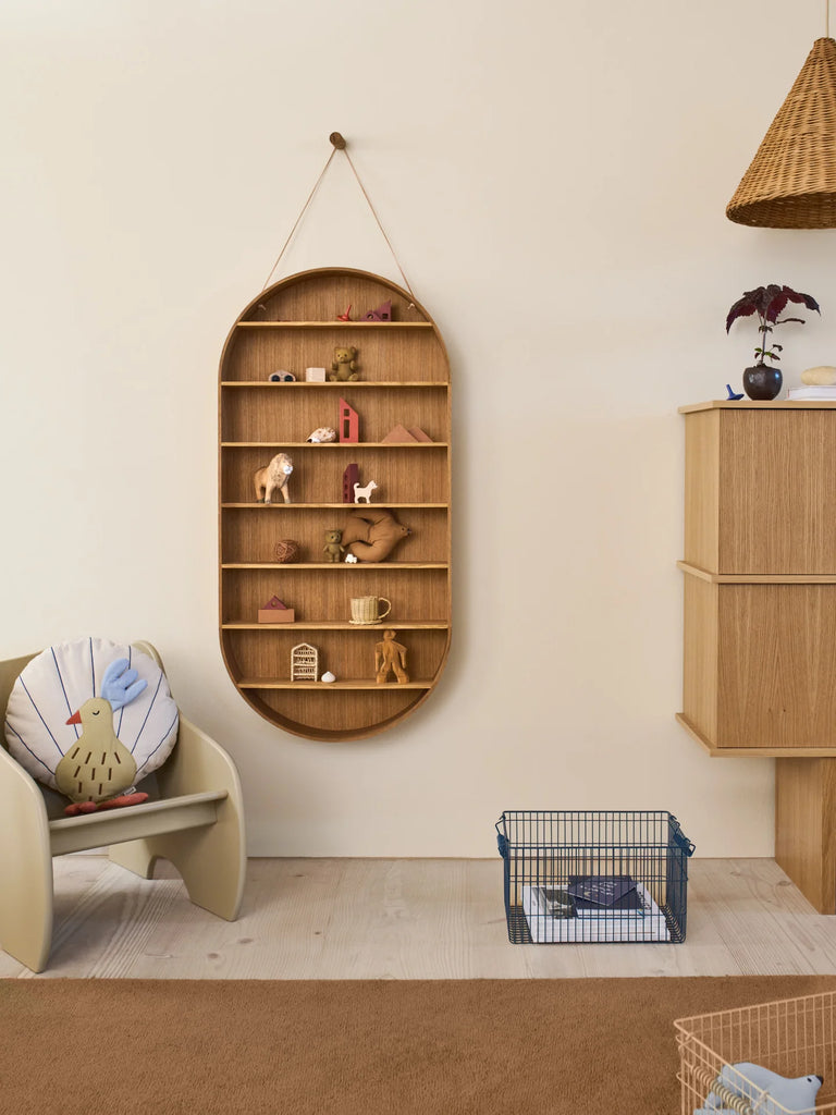 A cozy room features a minimalist Ferm Living Oval Oak Dorm Shelf displaying small toys and figurines, a beige chair with a bird pillow, wicker lampshade, small cabinet with a plant, and a blue wire pet cage on the floor.