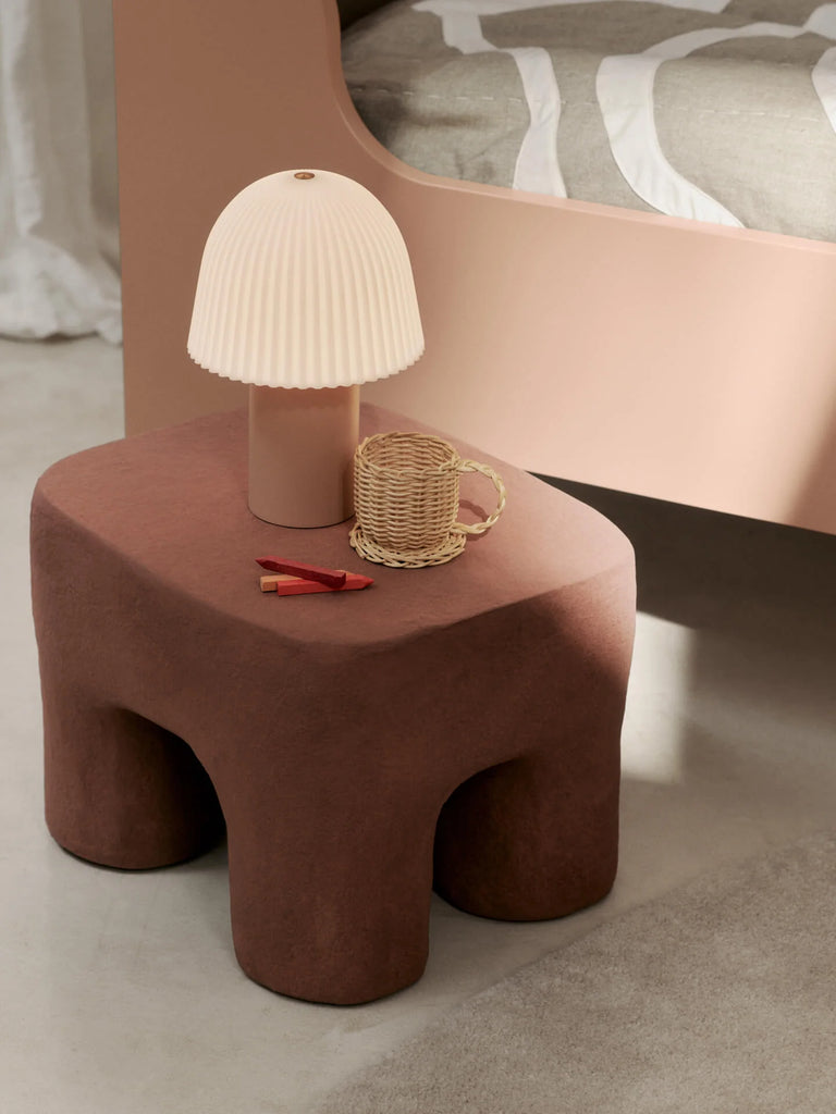 A modern bedside scene featuring the Ferm Living Frill Portable Lamp with a pleated beige shade on a sculptural table, alongside a woven rattan mug and small red decor, next to a bed dressed in neutral bedding.