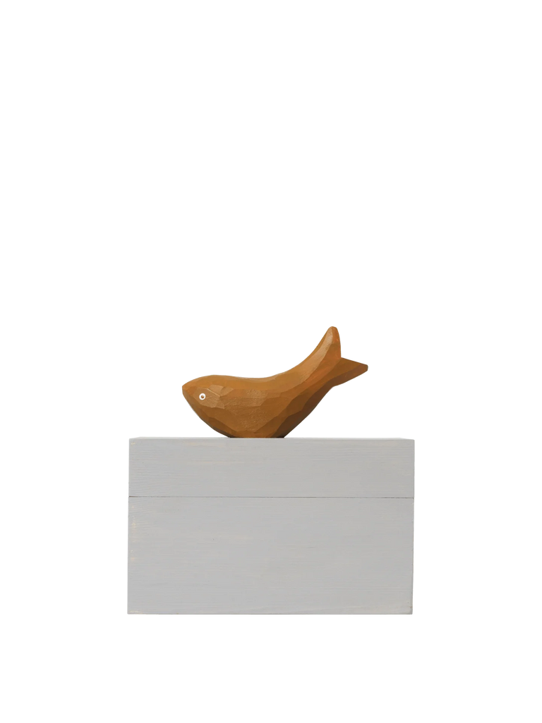 The Ferm Living Hand Carved Wooden Harbour Box - Fish features a small, fish-shaped wooden figure with a hand-carved handle resting on a rectangular light gray block, set against a plain black background.