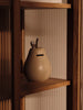The Ferm Living Pear Money Bank, a ceramic piggy bank with a coin slot, sits on a wooden shelf in warm lighting against vertical textured panels—a charming decor piece for any children's room.