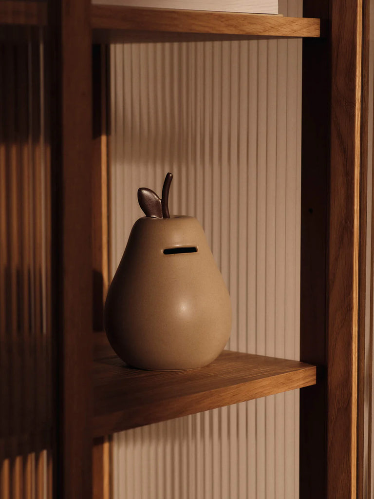 The Ferm Living Pear Money Bank, a ceramic piggy bank with a coin slot, sits on a wooden shelf in warm lighting against vertical textured panels—a charming decor piece for any children's room.